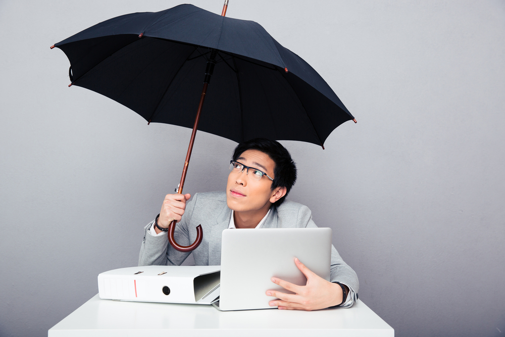 Young asian businessman sitting at the table with laptop and holding umbrella over gray background. Looking up